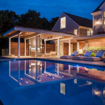 Modern home with covered outdoor living area, illuminated pool, and lounge seating at dusk