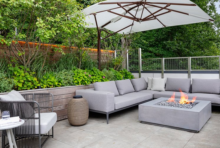Modern outdoor patio with gray sectional seating, a square fire pit, umbrella, and lush greenery in the background.